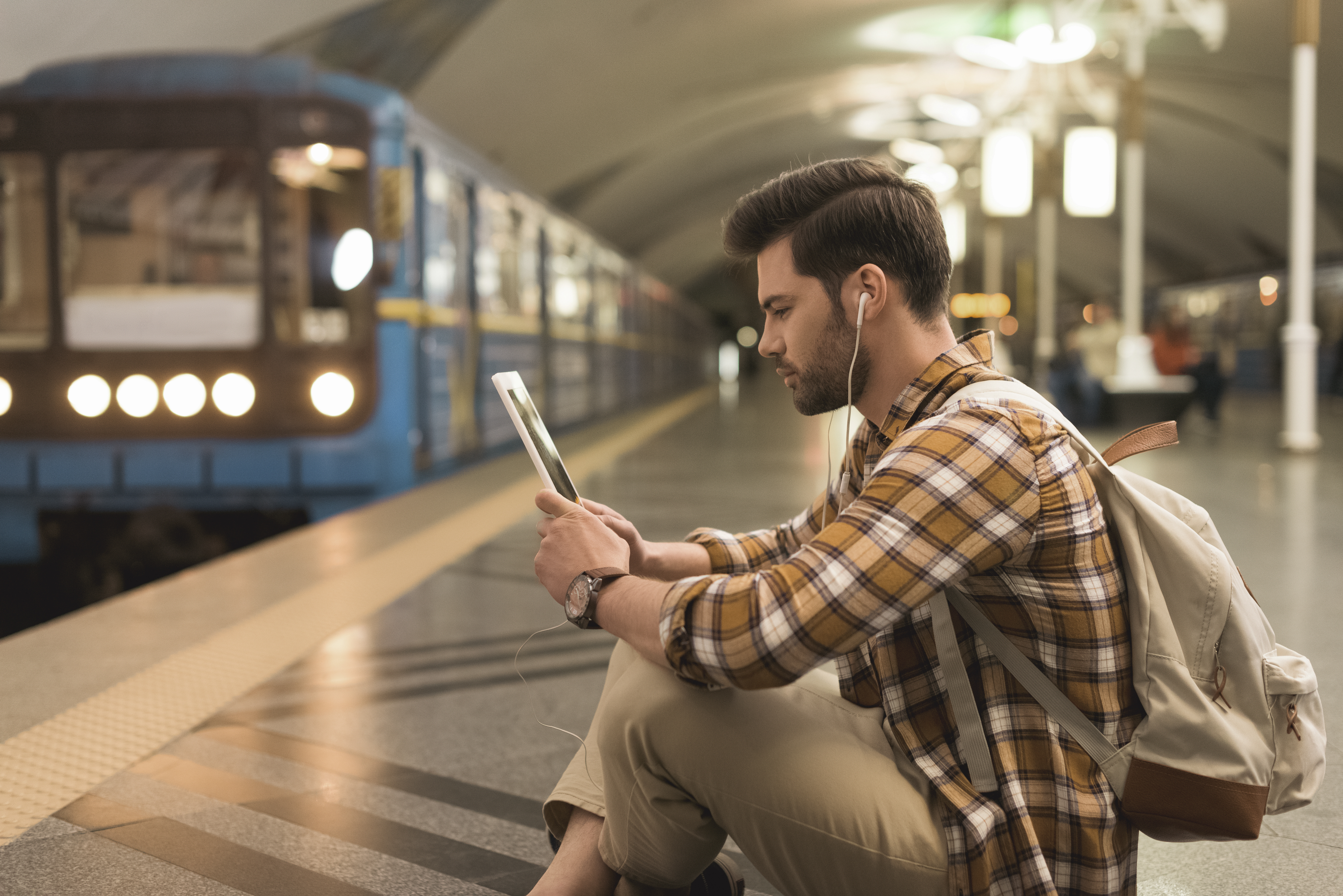 side view of young man with digital tablet and earphones sitting on floor at subway station 